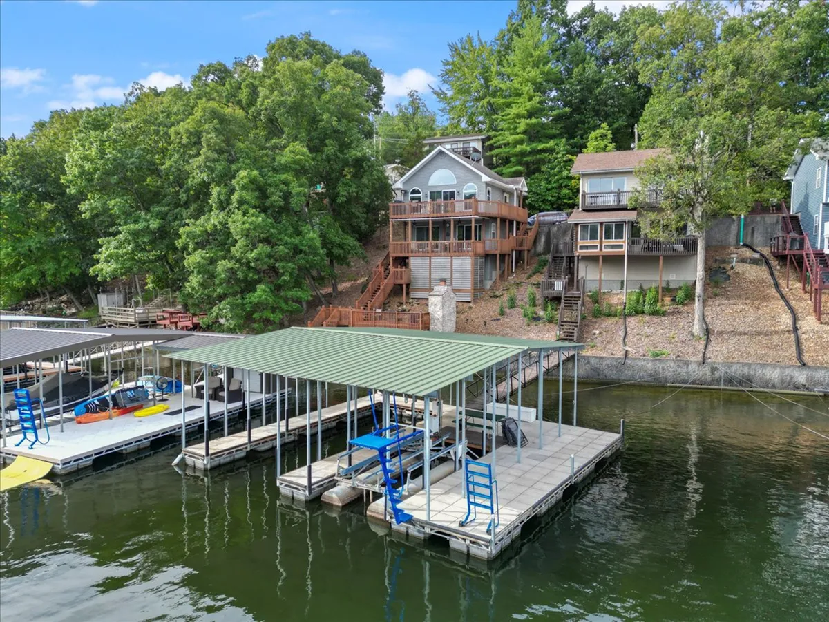 The image showcases a stunning lakefront scene featuring a private dock with boats ready for adventure, inviting wooden decks on the house overlooking the tranquil water, and lush greenery surrounding the property. The golden glow of sunset enhances the serene atmosphere, while the expansive outdoor spaces promise relaxation and fun.