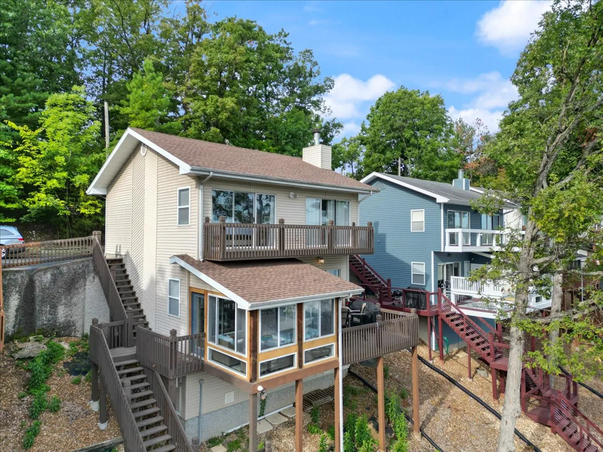 This image presents the exterior of a two-story house featuring a spacious deck surrounded by trees and neighboring houses. The deck suggests ample space for gatherings and outdoor fun, while the setting is bright, cheerful, and inviting.