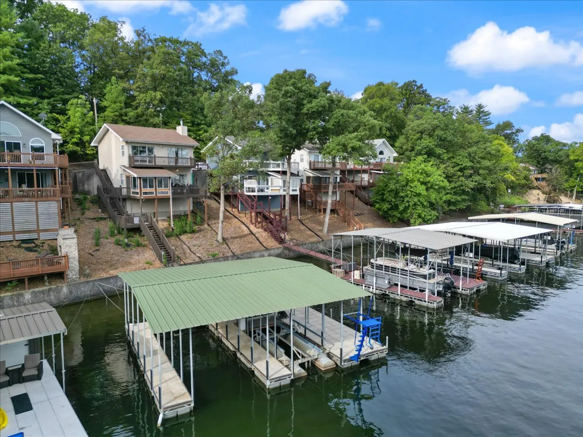 A scenic exterior shot featuring waterfront docks, houses nestled on a hillside, and lush greenery stretching down to the water. The tranquil lake setting is perfect for boating, fishing, and relaxing by the water's edge. The image conveys a serene, resort-inspired environment just steps from the home.