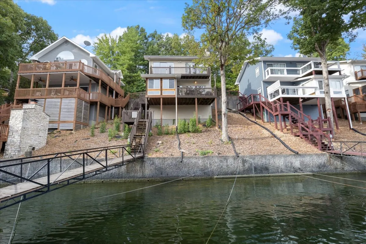 An exterior view featuring three distinct homes connected by wooden walkways, set amongst lush trees with the lake in sight. The scene conveys possibilities for outdoor activities like boating or family gatherings, and hints at nearby resort-style amenities.