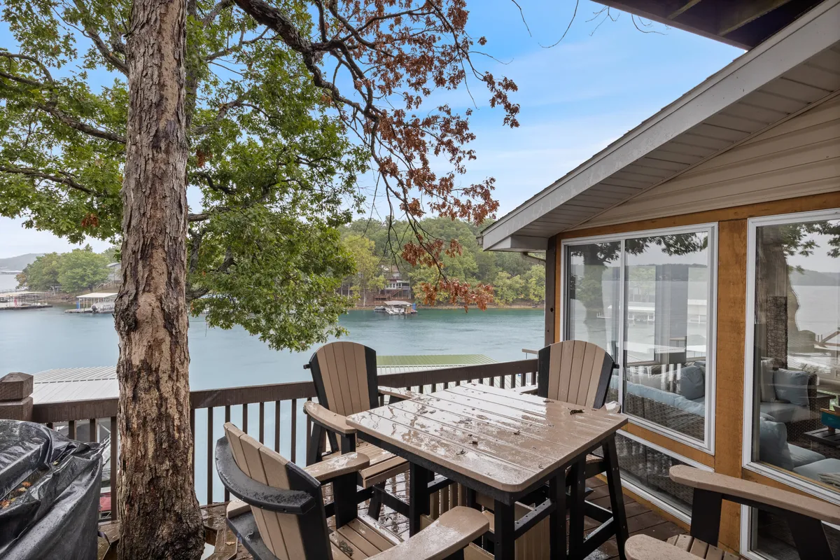 A spacious outdoor dining area on a wooden deck overlooks the serene lake, surrounded by trees. The deck features a large table and chairs, perfect for relaxing or socializing while enjoying the water views. The sunlight casts a warm glow, inviting residents to start or end the day immersed in nature.