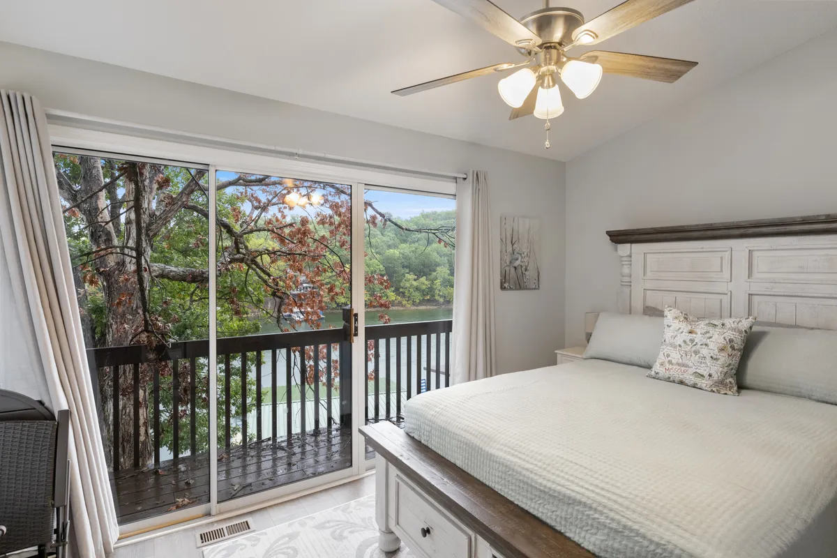 A bright bedroom with a bed positioned to maximize a balcony view, a prominent ceiling fan overhead, a decorative art piece, and gentle natural light streaming in. The space exudes comfort and a touch of whimsy, perfect for restful nights and inspired mornings.