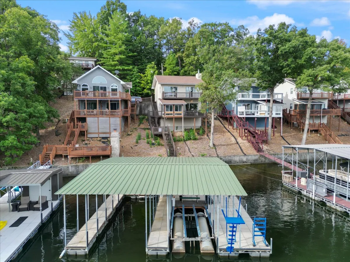 A peaceful scene of lakefront houses nestled among green trees, with a prominent dock featuring multiple boat slips extending into the water. The dock is large enough for boats and offers areas for lounging, with the calm lake reflecting the vibrant sky.