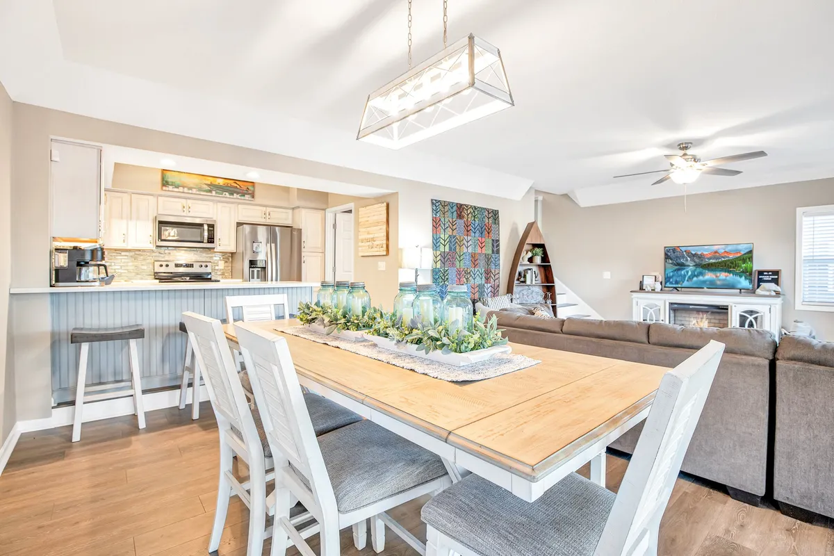 A spacious open-concept area with a dining table elegantly set with a centerpiece, a modern kitchen in the background, and a comfortable living room seating arrangement accented by decorative wall art. The scene is warm and inviting, perfect for family meals and social gatherings.