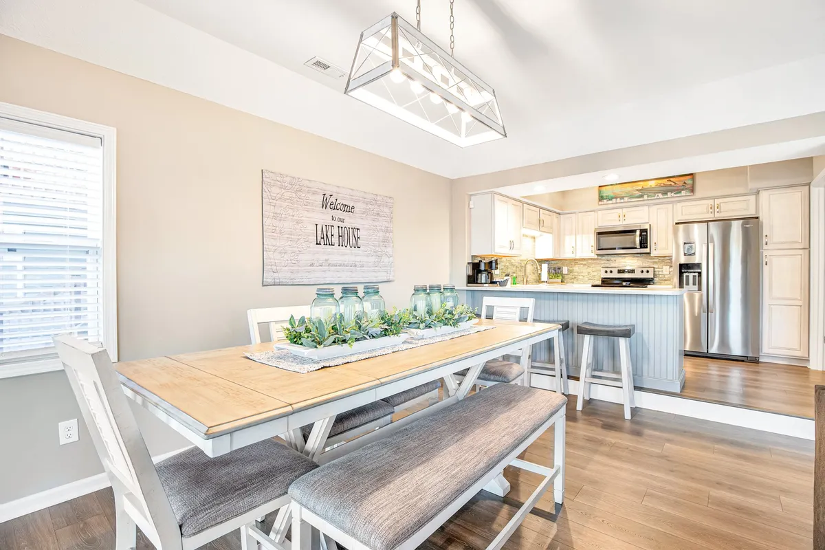 A modern dining table set for a gathering, with a charming 'welcome' sign visible in the background and an open kitchen area adjacent. The space is warm and inviting, suggesting thoughtful hospitality and the perfect setting for group meals or celebrations.